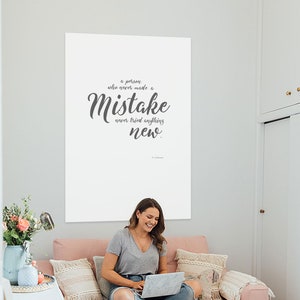 May include: A white poster with a quote in grey text that reads "A person who never made a mistake never tried anything new." The poster is hanging on a white wall in a room with a pink sofa and a woman sitting on it using a laptop.