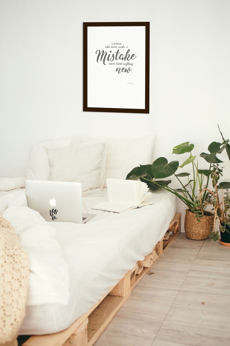 May include: A framed print with a white background and black text that reads "A person who never made a mistake never tried anything new." The print is hanging on a white wall above a bed with white bedding and a laptop computer on the bed.