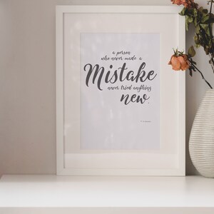 May include: A white framed print with a quote that reads "A person who never made a mistake never tried anything new." The quote is in a black cursive font. The print is sitting on a white table with a vase of dried flowers.