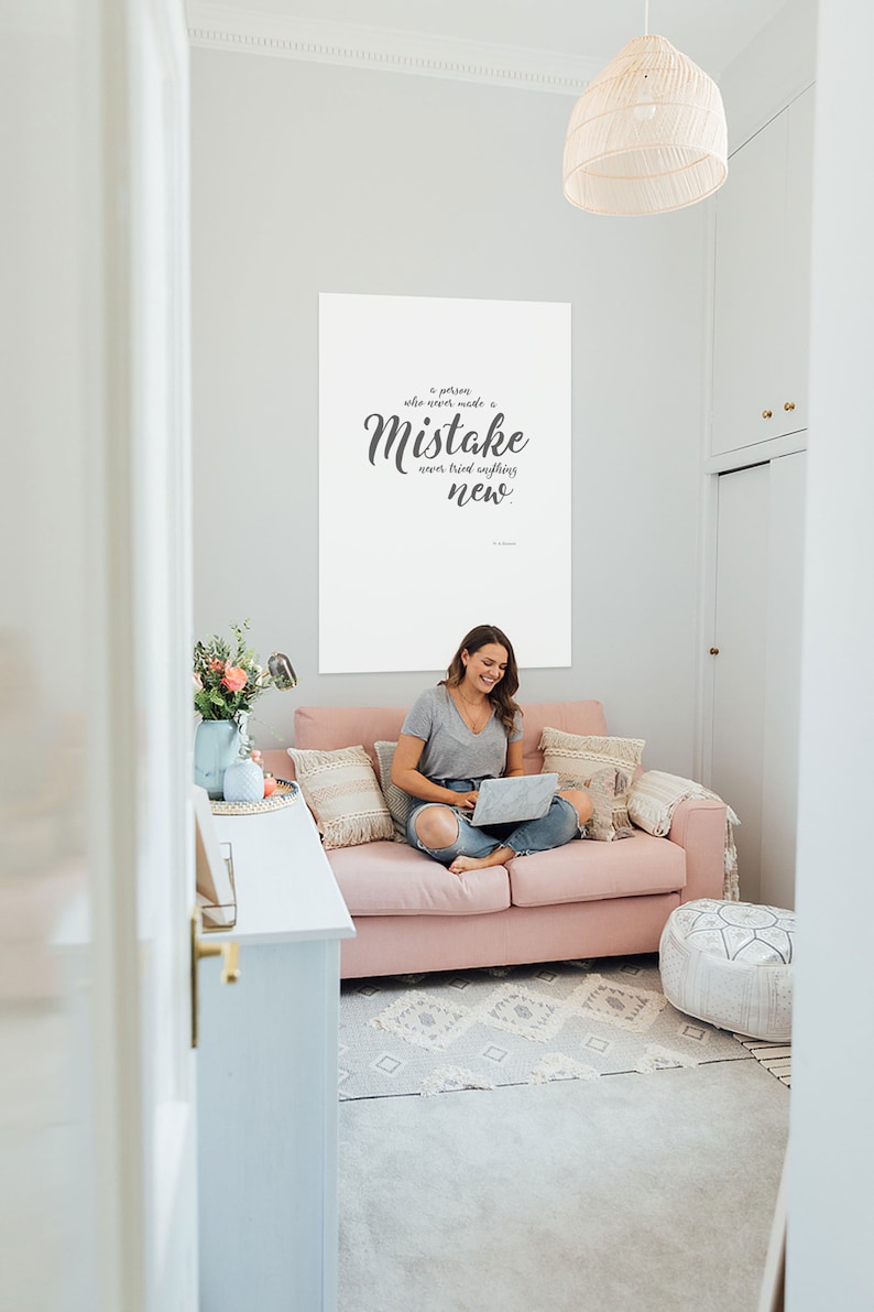 May include: A white poster with a quote in grey text that reads "A person who never made a mistake never tried anything new." The poster is hanging on a white wall in a room with a pink sofa and a woman sitting on it using a laptop.
