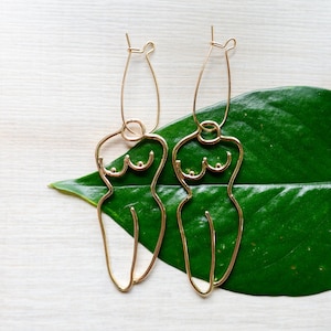以下が含まれることがあります： スタイリッシュな人間のシルエットの形をした、ゴールドトーンのイヤリングのペア。イヤリングは緑の葉からぶら下がっています。