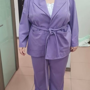 K&ouml;nnte beinhalten: Ein Anzug in Lavendelfarbe mit einer lang&auml;rmeligen Jacke und einer passenden Hose. Die Jacke hat einen breiten Kragen und einen Bindeg&uuml;rtel. Die Hose hat eine gerade Beinform.