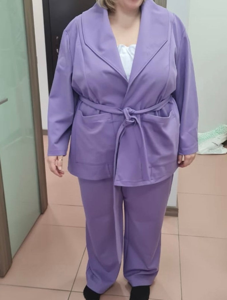 K&ouml;nnte beinhalten: Ein Anzug in Lavendelfarbe mit einer lang&auml;rmeligen Jacke und einer passenden Hose. Die Jacke hat einen breiten Kragen und einen Bindeg&uuml;rtel. Die Hose hat eine gerade Beinform.