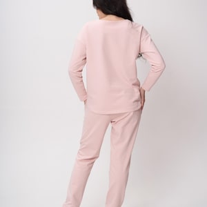 May include: A light pink long-sleeved top and matching long pants set. The top features a rounded neckline and long sleeves, while the pants have a straight-leg cut. The outfit is a solid, uniform pastel pink color throughout, paired with white sneakers.