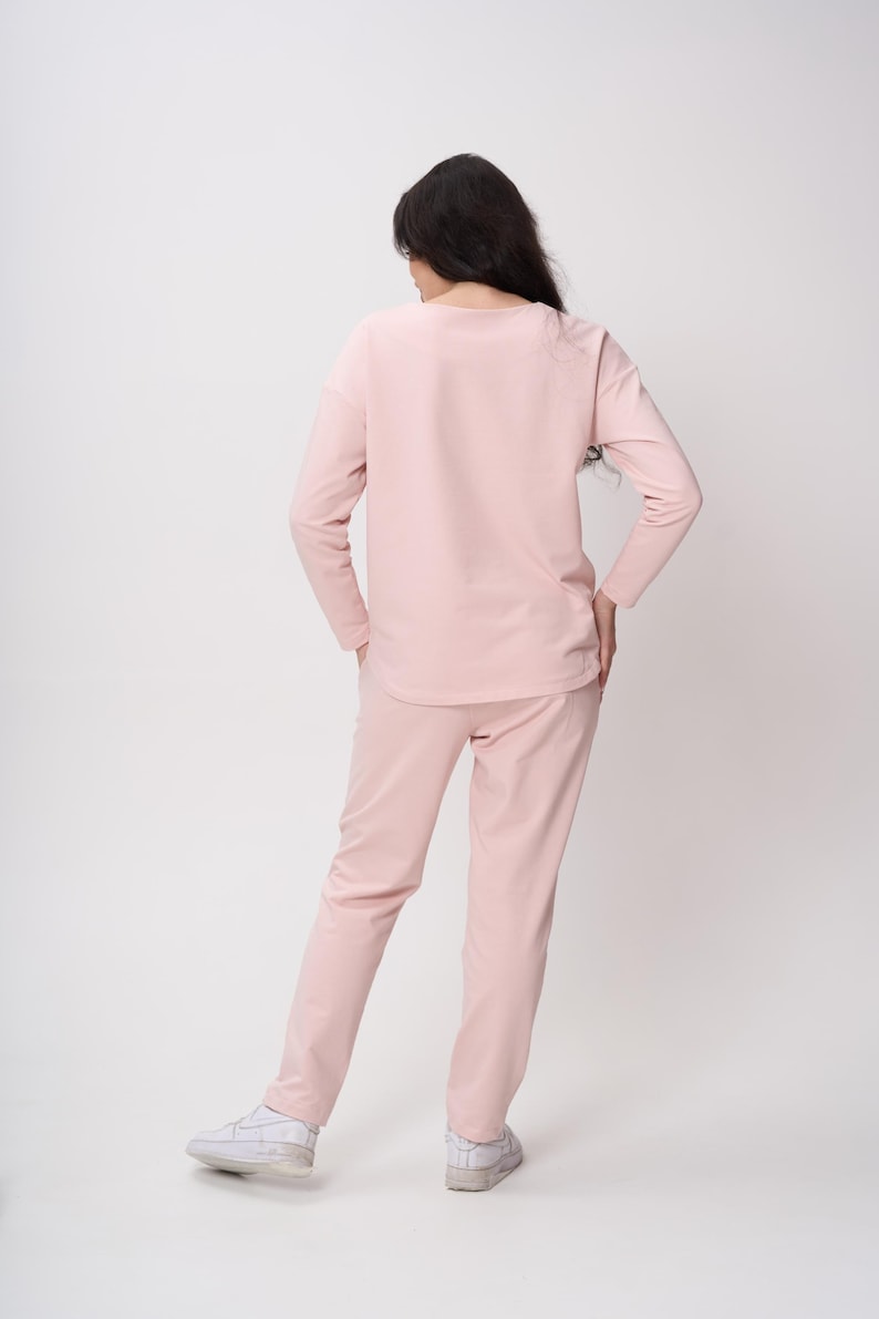 May include: A light pink long-sleeved top and matching long pants set. The top features a rounded neckline and long sleeves, while the pants have a straight-leg cut. The outfit is a solid, uniform pastel pink color throughout, paired with white sneakers.
