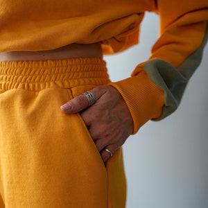 May include: A mustard yellow cropped sweatshirt and matching sweatpants with a grey detail on the sleeve. The person is wearing multiple silver rings on their finger.