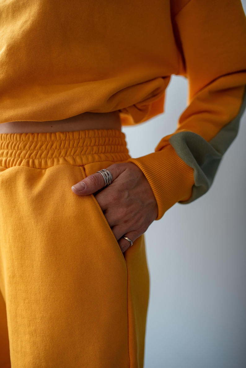 May include: A mustard yellow cropped sweatshirt and matching sweatpants with a grey detail on the sleeve. The person is wearing multiple silver rings on their finger.