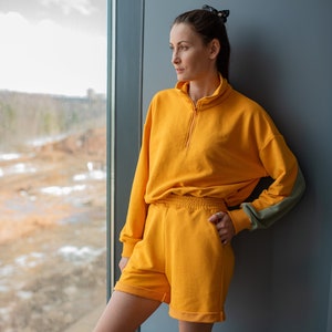 May include: A woman wearing a yellow tracksuit with a half-zip sweatshirt and matching shorts. She is standing by a window and looking out.