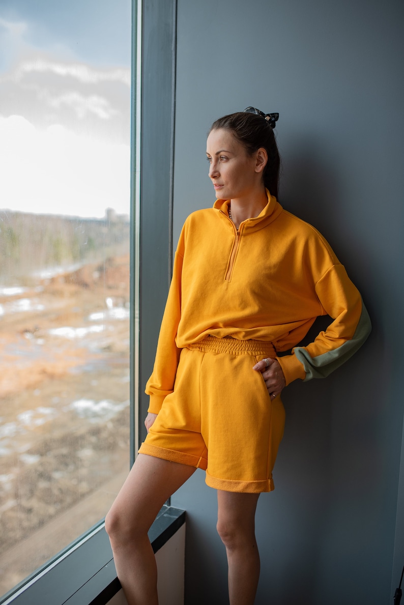 May include: A woman wearing a yellow tracksuit with a half-zip sweatshirt and matching shorts. She is standing by a window and looking out.