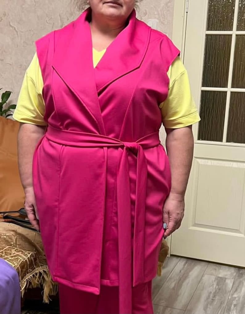 K&ouml;nnte beinhalten: Eine rosa &auml;rmellose Weste mit einem breiten Kragen und einem passenden G&uuml;rtel. Die Weste ist aus einem weichen, dehnbaren Stoff gefertigt und hat eine lockere Passform.
