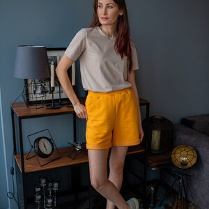 May include: A woman wearing a light beige t-shirt and bright yellow shorts stands in front of a shelf with a lamp and clock. She is wearing gray sneakers.