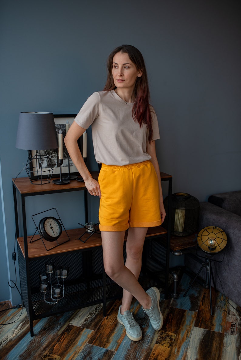 May include: A woman wearing a light beige t-shirt and bright yellow shorts stands in front of a shelf with a lamp and clock. She is wearing gray sneakers.