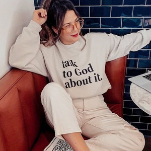May include: A woman wearing a white sweatshirt with the text "talk to God about it." She is sitting on a brown leather couch with a laptop on a marble table beside her. She is wearing white jeans and leopard print high-top sneakers.