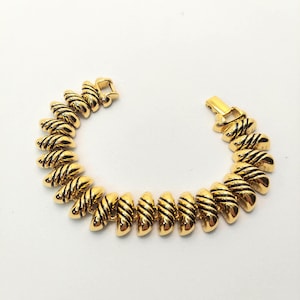 May include: Gold-toned bracelet with a repeating design of curved, ridged segments. The bracelet has a clasp closure. The segments are a shiny gold color with black detailing. The jewelry is displayed on a white background.