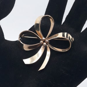 May include: A gold-toned bow brooch with a pin closure. The brooch features a three-dimensional bow design with rounded loops and pointed ends. The center of the bow has three small, round embellishments. The brooch is displayed on a black surface.