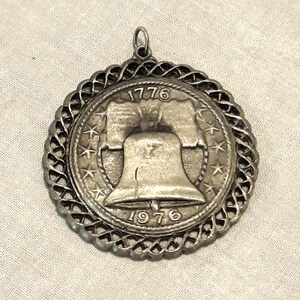 May include: Silver-toned pendant featuring a Liberty Bell design with the years 1776 and 1976 inscribed. The pendant is surrounded by a decorative border of stars and a rope-like design.