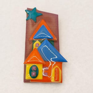May include: A colorful ceramic house brooch with a blue roof, orange walls, and a yellow door. A green star is on top of the house.