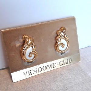 May include: A pair of gold-tone clip-on earrings with white enamel accents. The earrings are in the shape of a stylized swirl and are packaged on a card that reads "VENDOME-CLIP".