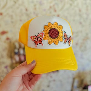 May include: Yellow and white trucker hat with a sunflower and butterfly design. The hat has a yellow peak and mesh back, with a white front panel displaying a sunflower and two butterflies. The design is embroidered.