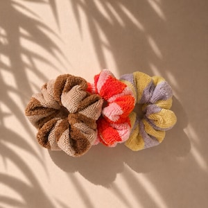 Peut inclure: Trois chouchous circulaires fabriqués dans un tissu texturé semblable à une serviette. Le premier chouchou présente des rayures alternées marron et beige, le deuxième des rayures rouge vif et rose, et le troisième des rayures violet clair et jaune.