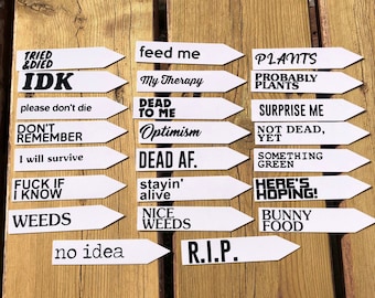 May include: A selection of white plant markers with black text, arranged on a wooden surface. The markers feature humorous phrases such as "Feed Me," "Weeds," and "Here's Hoping!" Ideal for marking plants in a garden.
