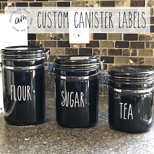 Op de afbeelding: Drie zwarte potten met zilveren accenten en scharnierende deksels. De potten zijn gelabeld met "FLOUR", "SUGAR" en "TEA" in witte letters. De afbeelding bevat ook de tekst "CUSTOM CANISTER LABELS".