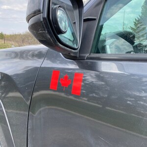 May include: A close-up of a dark gray vehicle's side panel with a red Canadian flag sticker. The flag features a red maple leaf in the center and two red vertical bars on either side. The side mirror is visible.