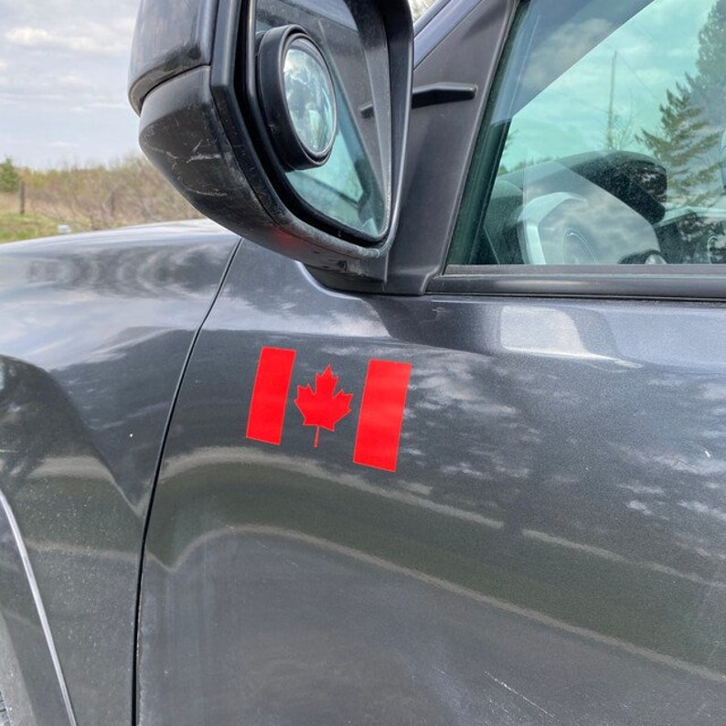 May include: A close-up of a dark gray vehicle's side panel with a red Canadian flag sticker. The flag features a red maple leaf in the center and two red vertical bars on either side. The side mirror is visible.