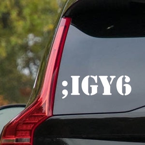 Könnte beinhalten: Weißes Vinyl-Aufkleber mit dem Text ";IGYG" auf einer Autoscheibe. Der Aufkleber ist in einer fetten, serifenlosen Schriftart. Das Rücklicht des Autos ist rot und das Auto ist schwarz.