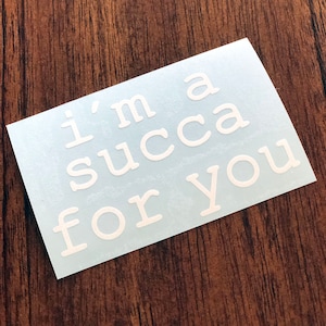 May include: A white sticker with the text "I'm a succa for you" in a rounded font. The sticker is on a light blue background, placed on a dark wood grain surface. A fun, quirky item.
