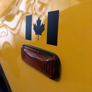 May include: A close-up of a yellow vehicle with a black Canadian flag decal and a red side marker light. The flag features a black maple leaf and two black rectangles. The side marker light has a black border.