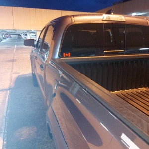 May include: A gray pickup truck with a black bed and a Canadian flag sticker on the rear window. The truck is parked outdoors at dusk or night, with a bridge and other vehicles visible in the background.