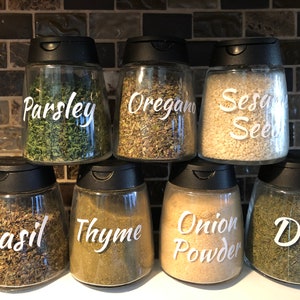May include: Seven clear spice jars with black lids are arranged on a shelf. Each jar is labeled with white cursive text indicating the contents: parsley, oregano, sesame seeds, basil, thyme, onion powder, and dill. The jars are filled with various dried herbs and spices.