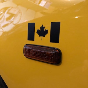 May include: A close-up of a yellow surface with a black Canadian flag decal and a rectangular amber light. The flag features a black maple leaf in the center and two black vertical bars on either side.