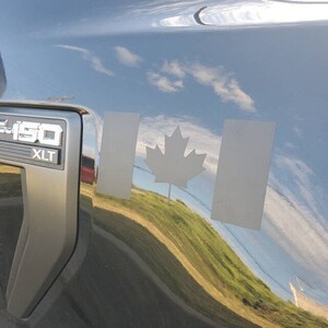 May include: A black vehicle with a silver F-150 XLT emblem. A gray Canadian flag decal is on the vehicle's body, featuring a maple leaf and two vertical rectangles. The vehicle's surface reflects a sky with clouds and a green hill.