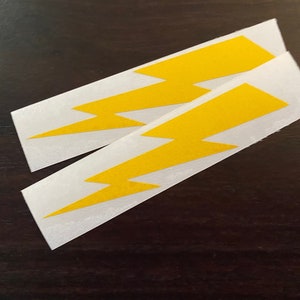 May include: Two yellow lightning bolt stickers on a white background. The stickers are rectangular with a yellow lightning bolt design. The stickers are on a dark brown surface.