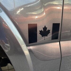 May include: A black decal of the Canadian flag, featuring a maple leaf and two vertical bars, is affixed to a gray vehicle. The decal is positioned on the side of the vehicle, near the door.