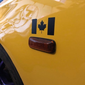 May include: A close-up of a yellow car panel with a black Canadian flag decal and a rectangular amber side marker light. The flag features a black maple leaf in the center.