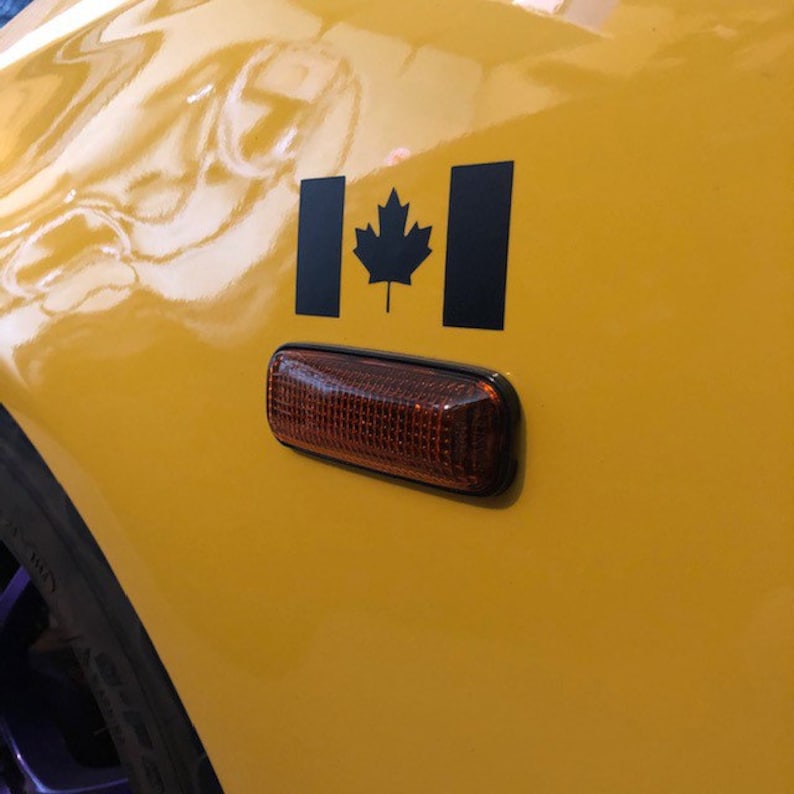 May include: A close-up of a yellow car panel with a black Canadian flag decal and a rectangular amber side marker light. The flag features a black maple leaf in the center.