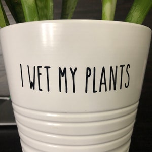 May include: White ceramic plant pot with horizontal ridged detailing. The text "I WET MY PLANTS" is printed in black, centered on the pot. Green and white striped plant leaves are visible at the top of the pot.