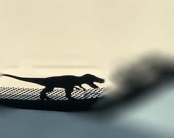Könnte beinhalten: Schwarze Silhouette eines Tyrannosaurus Rex-Dinosauriers mit offenem Maul, vor hellem Hintergrund. Der Dinosaurier befindet sich auf einer perforierten Oberfläche, mit einer verschwommenen, dunklen Form rechts.