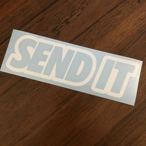 Op de afbeelding: Een witte rechthoekige sticker met de woorden "SEND IT" in een vet, afgerond lettertype. De tekst is wit omlijnd en geplaatst tegen een lichtblauwe achtergrond. De sticker ligt op een houten oppervlak.