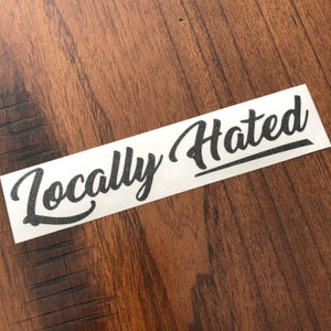 Puede incluir: Una pegatina blanca con las palabras "Locally Hated" en fuente de escritura negra. La pegatina es rectangular y está colocada sobre una superficie de madera marrón.