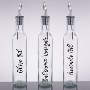 May include: Three clear glass bottles with stainless steel pour spouts. Each bottle has a black label with the words "Olive Oil", "Balsamic Vinegar", and "Avocado Oil" in a cursive font. The bottles are arranged in a row against a light background.