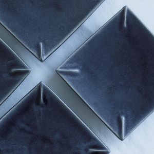 May include: Four square, dark blue ceramic plates arranged diagonally. Each plate has a slightly raised rim and a small indentation at each corner. The plates are on a white surface, creating a minimalist aesthetic.