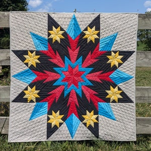 Puede incluir: Una colcha con un gran diseño de estrella de múltiples puntas en tonos de rojo, azul, negro y amarillo. La estrella está sobre un fondo gris claro.