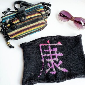 May include: A black knitted headband with a purple and pink kanji character embroidered on it. The character is "異" which means "different" or "strange".