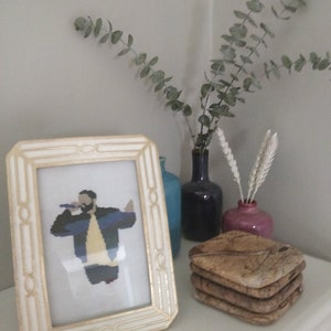 Peut inclure: Un cadre blanc avec une bordure dorée entoure une broderie au point de croix d'une personne chantant dans un microphone. La broderie est sur un tissu blanc et la personne porte une veste bleue, une chemise jaune et un pantalon noir. Le cadre est sur une surface blanche avec une pile de sous-verres en bois et un vase avec des fleurs séchées.