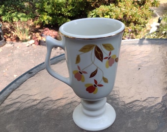 May include: A white pedestal coffee mug with a gold rim and handle. The mug is decorated with a floral pattern in shades of brown, yellow, and red. The mug is approximately 5 inches tall.