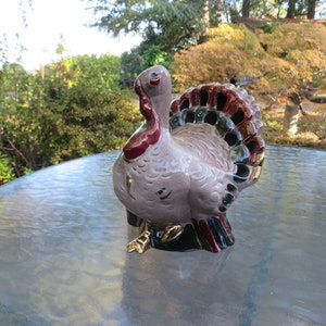 May include: A ceramic turkey figurine with a light pink body, a red wattle and head, and a fan of colorful feathers. The turkey has gold accents on its feet and is set on a glass table with a blurred outdoor background.